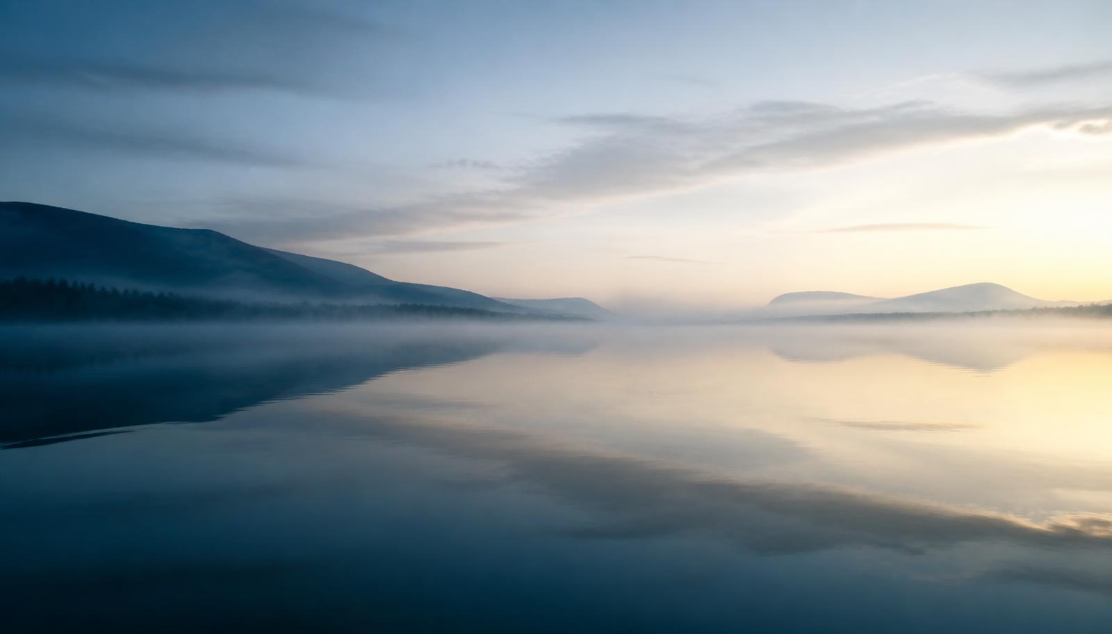 Still water at first light, with distant hills emerging through low mist.