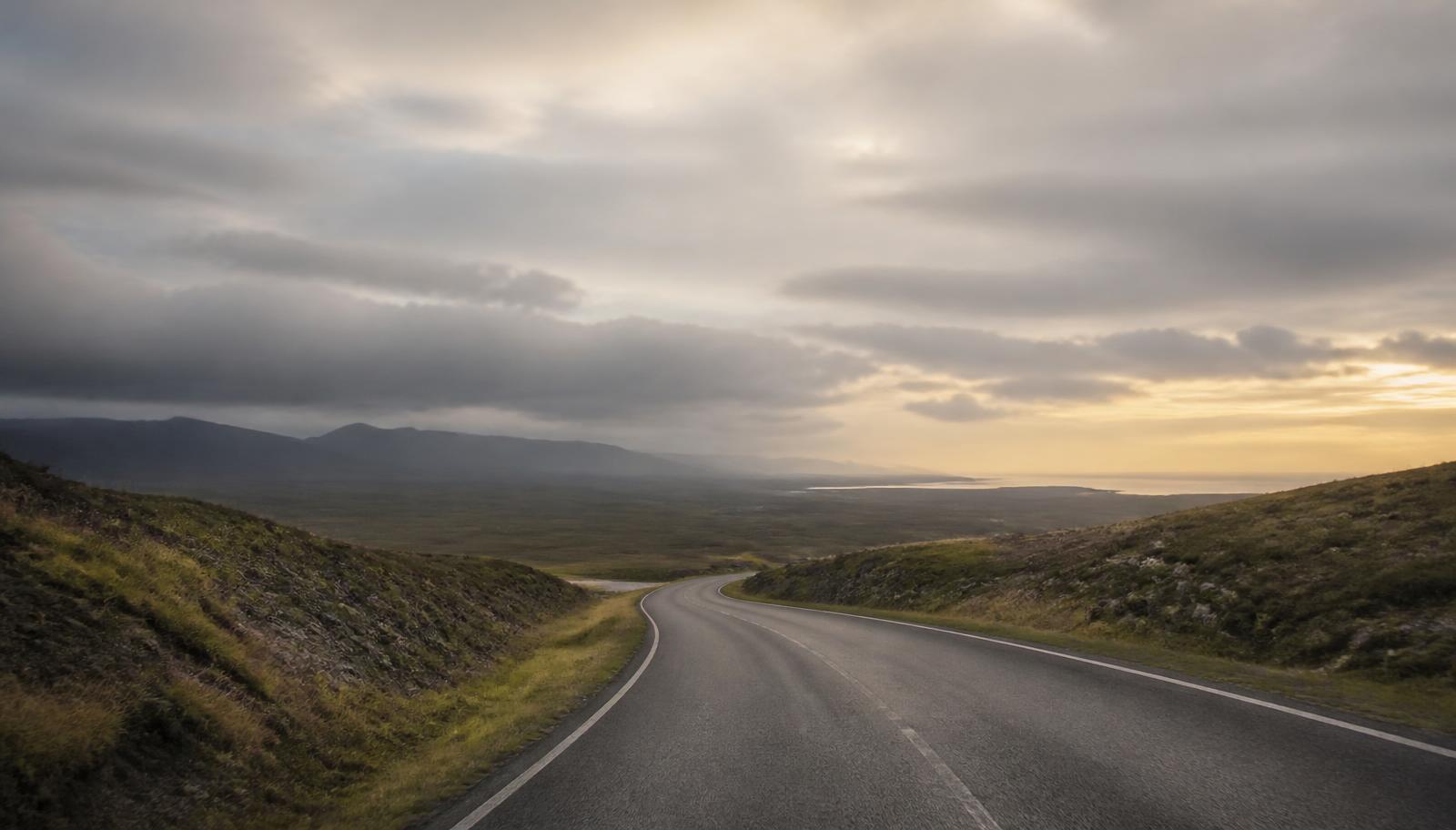 An empty road curving through open country toward a distant, brightening horizon.